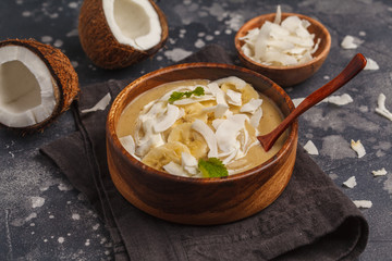 Coconut banana smoothie bowl in a wooden plate on a dark background, vegan healthy food concept. Top view, copy space.