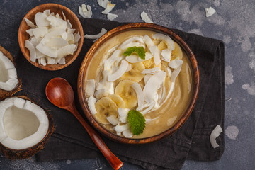 Coconut banana smoothie bowl in a wooden plate on a dark background, vegan healthy food concept. Top view, copy space.