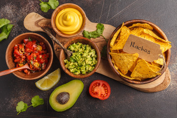 Mexican food concept. Nachos - yellow corn totopos chips with various sauces in wooden bowls: guacamole, cheese sauce, pico del gallo. Top view, food background