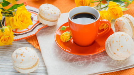 Orange cup of black coffee, yellow roses flowers and sweet pastel french macaroons