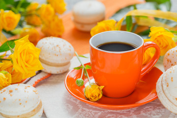 Orange cup of black coffee, yellow roses flowers and sweet pastel french macaroons