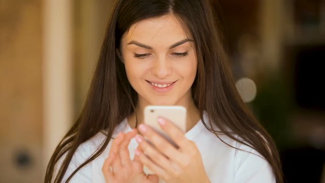 Intrigued girl smiles for the camera while texting on the phone, inloved caucasian woman using gadget in cozy brown room