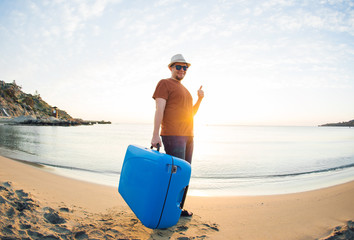 Man holding suitcase and showing thumbs up against the blue ocean. Travel concept.
