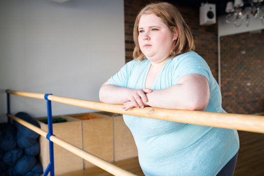 Waist up portrait of obese young woman standing at bar in dance class looking pensively at window, copy space