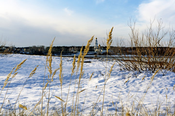 dry golden spikelets of last year's grass against the background of a blurry spring river landscape with an orthodox church on the far shore on a sunny day