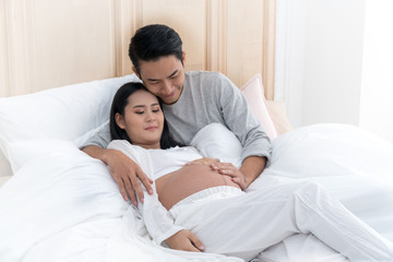 Fototapeta premium Mąż z miłością obejmujący brzuch żony na białym łóżku. Szczęśliwa para młody człowiek przytula swoją ciężarną piękną żonę w sypialni.