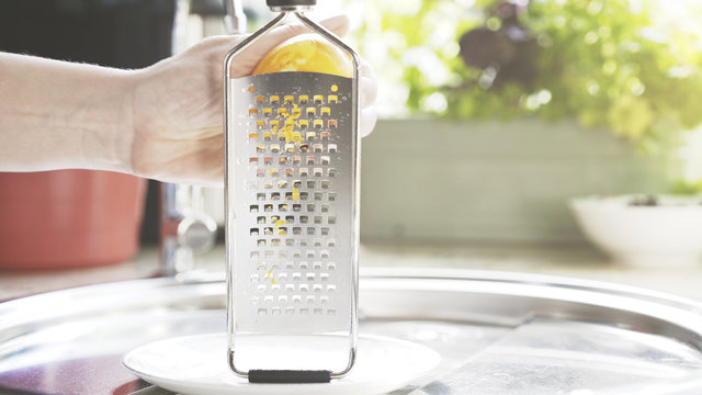 A young woman is holding a grater and peel a lemon zest into a small white plate.