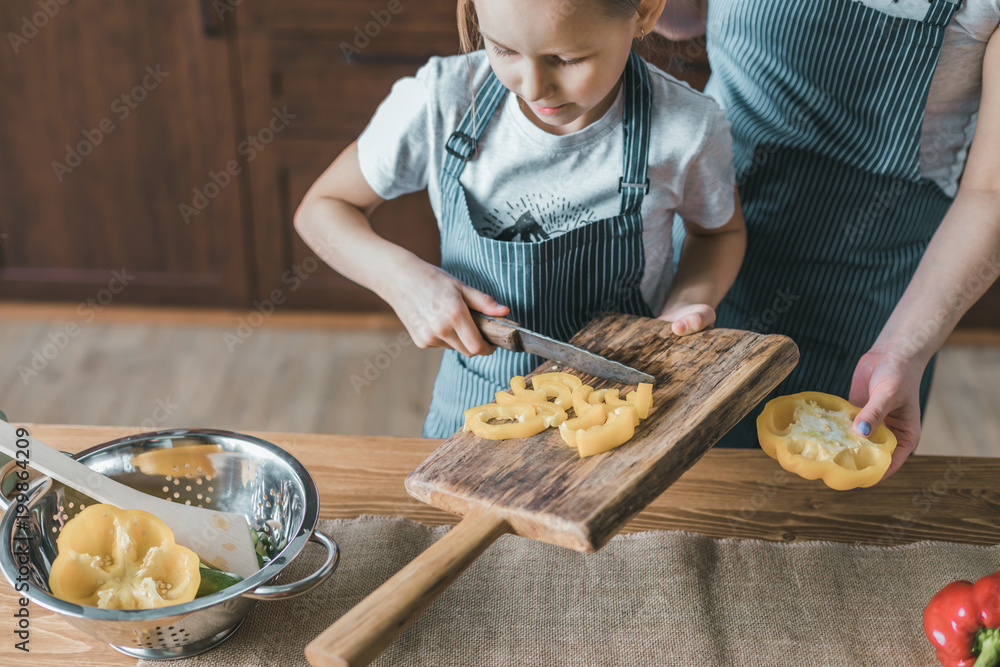 Canvas Prints Young girl putting cut pepper to bowl while cooking with mother.