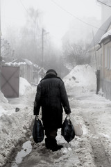 Rear view sad homeless old woman in dirty clothes with bags walks along street of fogy winter city
