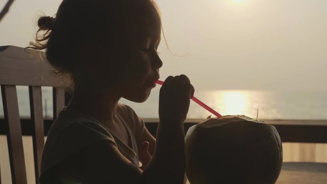 Child girl sits at seaview cafe and drinks coconat water in slow motion