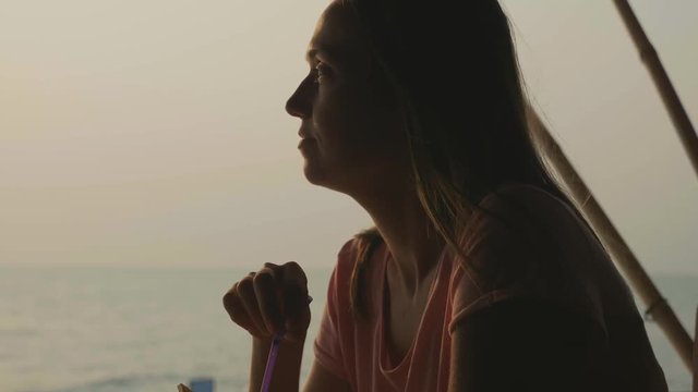 Young woman sits at seaview cafe and drinks coconat water in slow motion
