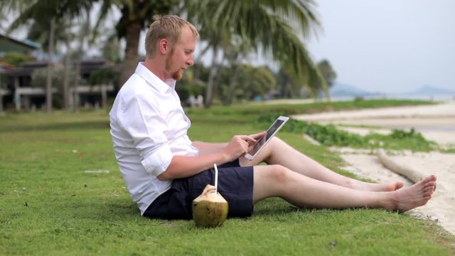 man with a tablet tablet and drinks coconut on the beach