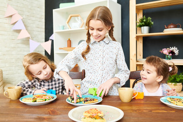 Smiling little girl with two braids standing at wooden table of spacious dining room and playing with homemade waffles and cookie cutter, her sisters keeping eye on her