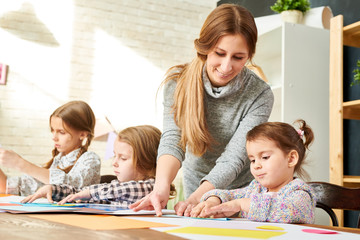 Working process at art studio: little pupils sitting at wooden desk and wrapped up in greeting cards creation from colored paper while their friendly young teacher assisting them