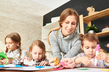Obraz premium Concentrated little girls sitting at wooden desk of modern art studio and expressing creativity with help of colored paper while their friendly young teacher keeping eye on them.
