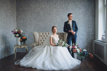 A beautiful bride is sitting in the studio on the couch in a lace dress. The groom looks out the...