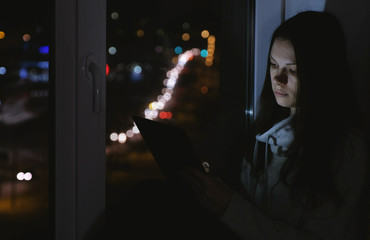 Woman sitting on the windowsill in the dark night and using tablet for web.