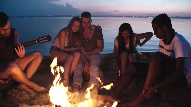 PAN of young people camping on beach at dusk: they warming up by fire as bearded man playing guitar