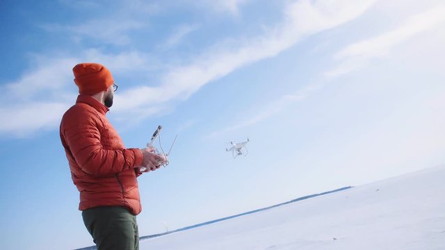 Man in a bright jacket against a clear blue sky and white snow launches a white drone for an aero photo. Stylish man controls a quadrocopter in the open space. Flight of a quadrocopter in winter.