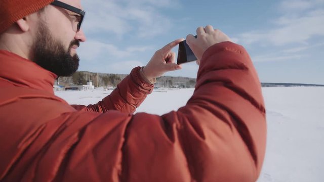 A traveler in a bright jacket stands against the sun and takes pictures of the winter nature on the smartphone. Hicker takes a photo on the phone: the admiration of nature on a sunny day.