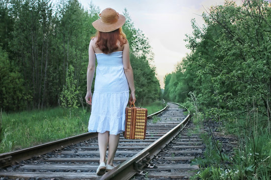 girl in a white sundress and wicker suitcase walking on rails