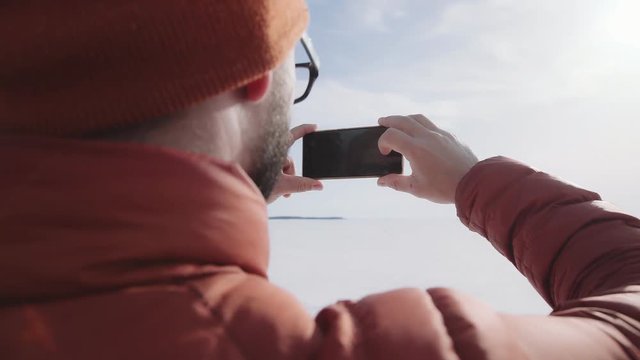 POV. A man with a beard and wearing glasses takes off on a smartphone on a sunny day. Hicker man taking a photo of winter nature. The tourist admires nature and makes photos.
