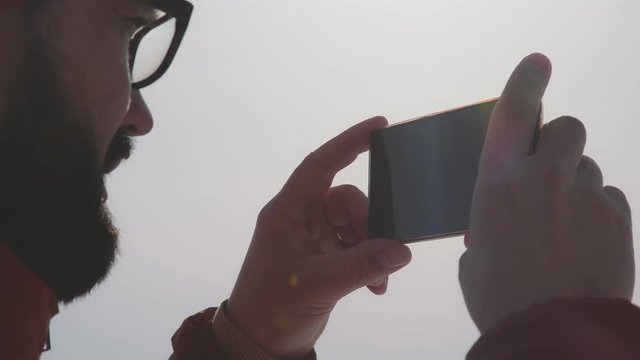 POV. A man with a beard and wearing glasses takes off on a smartphone on a sunny day. Hicker man taking a photo of winter nature. The tourist admires nature and makes photos.