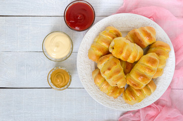 Soft homemade buns with sausage on a ceramic plate on the white wooden background with different sauces. Sausage in the dough. Top view.