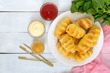 Soft homemade buns with sausage on a ceramic plate on the white wooden background with different sauces. Sausage in the dough. Top view.