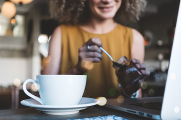 Handsome woman in city cafe, with cup of coffee and latop, adding sugar into her drink