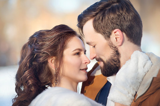 The bride in a fur coat and white mittens holds the groom by the neck and draws a close-up on a winter day.