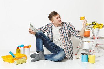 Young happy man holds bundle of dollars, cash money, cretit card, sits on floor with instruments...