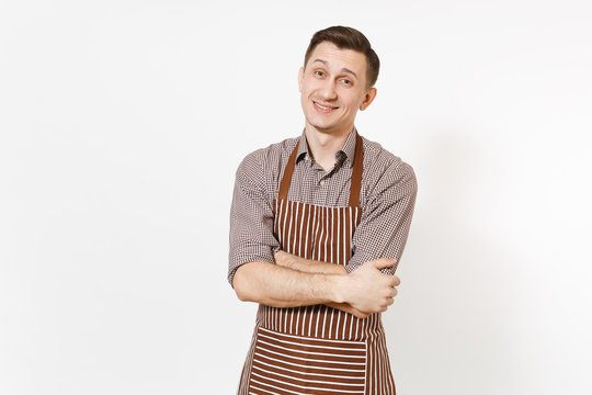 Young smiling man chef or waiter in striped brown apron, shirt holding hands folded isolated on white background. Male housekeeper or houseworker looking camera. Domestic worker for advertisement.