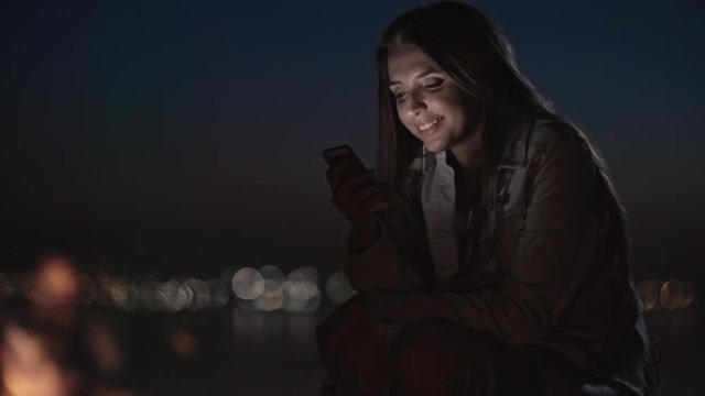 Tilt up of cheerful young woman with mobile phone laughing and sitting by campfire at night