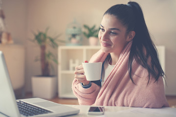 Cheerful girl at home sitting on floor holding cup of coffee and using laptop.