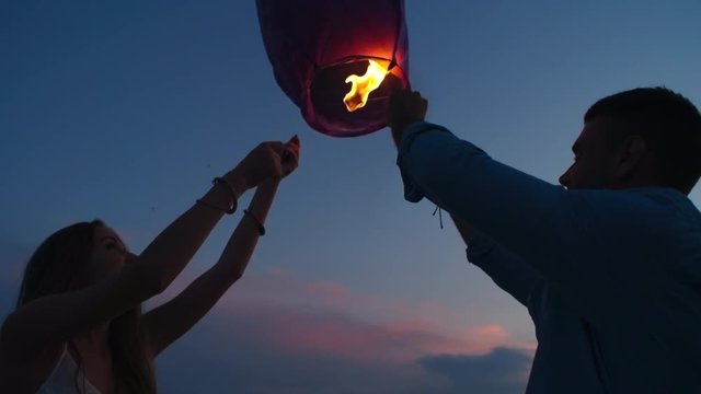 Tilt up of loving couple releasing Chinese lantern into sky on romantic date on beach at dusk