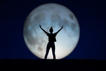 Fototapeta premium Silhouette of a woman standing in the night with the arms up, giant moon in the background