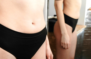 Thin young woman examines the folds on her stomach standing in front of a mirror in her underwear. Belly and arms close up.