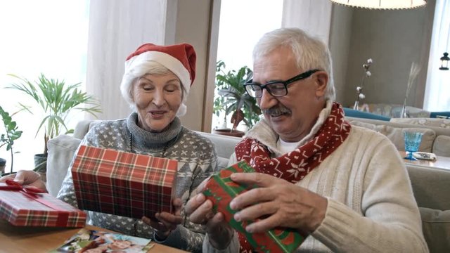 Surprised senior man and woman in Santa hat sitting on couch at restaurant table and opening Christmas gifts at holiday dinner
