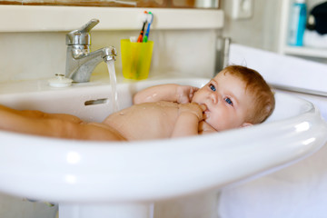 Cute adorable baby taking bath in washing sink and grab water tap.