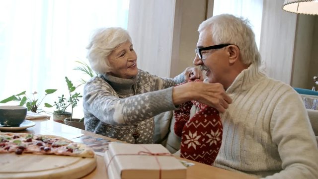 Senior woman taking knitted red scarf with snowflakes from gift box and putting it on husband while celebrating Christmas in restaurant