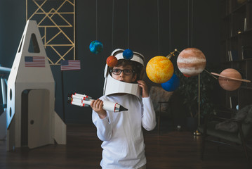 Cute little boy wearing cardboard astronaut helmet flying toy rocket through planets, cardboard spaceship rocket in the background © supamotion