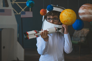 Cute little boy wearing cardboard astronaut helmet flying toy rocket through planets, cardboard...
