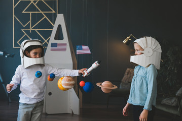 Cute little dreamer siblings playing with Solar System model at home, pretending to be astrounauts, cardboard space rocket in the background