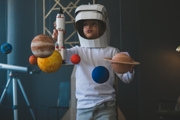Cute little boy wearing cardboard astronaut helmet flying toy rocket through planets, cardboard...