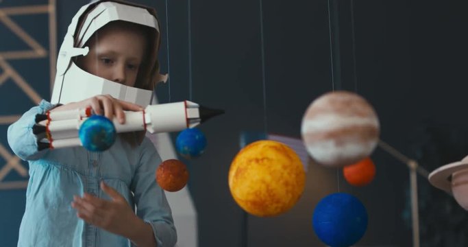 CU Cute little girl wearing cardboard astronaut helmet launching a toy rocket from a spaceport through planets. 4K UHD 60 FPS SLOW MO