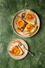 Homemade crepes pancakes served in ceramic plates with bloody oranges and rosemary syrup with sliced sicilian red oranges over green texture background. Top view, space