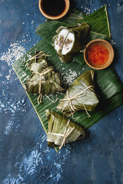 Asian rice piramidal steamed dumplings from rice tapioca flour with meat filling in banana leaves. Ingredients and sauces above over blue texture background. Top view, space.