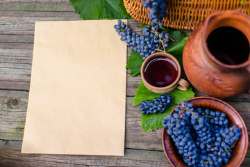 Sheet of paper for text lay beside basket,bowl with grapes, jar and cup with wine on rustic wood. Wine making background. Top view