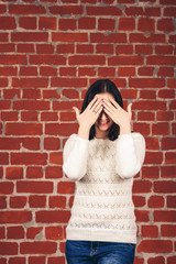 Beautiful girl in white sweater closes her eyes with her hands against backdrop of brick wall. Copy space.
